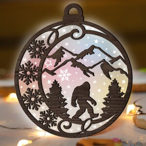 May include: A wooden Christmas bauble shaped like a circle with a cut-out design of a Bigfoot standing in a snowy mountain landscape. The bauble is painted with a gradient of pink, orange, and yellow colours, and features snowflakes and stars.