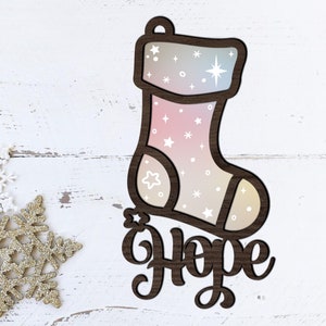 May include: A wooden Christmas stocking ornament with a pink and blue ombre design and white stars. The stocking has the word "Hope" written below it.