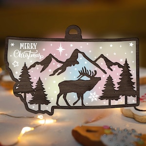 May include: A wooden Christmas ornament shaped like the state of Montana with a silhouette of an elk and mountains. The ornament has a rainbow-coloured background with white snowflakes and stars. The text "Merry Christmas" is written in white on the ornament.