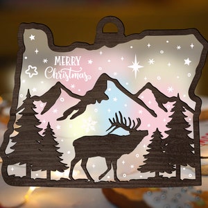 May include: A wooden ornament shaped like the state of Oregon with a silhouette of a mountain range, a deer, and two pine trees. The ornament is painted with a rainbow gradient and has the words "Merry Christmas" written on it.