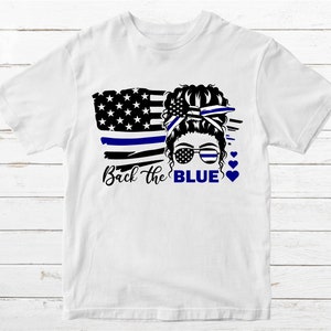 Puede incluir: Camiseta blanca con un gr&aacute;fico negro y azul de una mujer con un mo&ntilde;o en el pelo, que lleva gafas de sol y una cinta azul. El gr&aacute;fico tambi&eacute;n incluye una bandera estadounidense estilizada con una l&iacute;nea azul. El texto "Back the BLUE" est&aacute; impreso debajo del gr&aacute;fico.