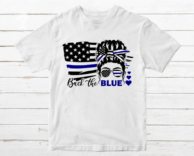 Puede incluir: Camiseta blanca con un gr&aacute;fico negro y azul de una mujer con un mo&ntilde;o en el pelo, que lleva gafas de sol y una cinta azul. El gr&aacute;fico tambi&eacute;n incluye una bandera estadounidense estilizada con una l&iacute;nea azul. El texto "Back the BLUE" est&aacute; impreso debajo del gr&aacute;fico.