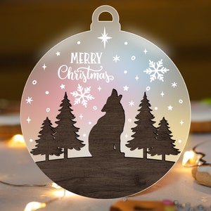 May include: A Christmas ornament with a wolf howling at the moon, surrounded by pine trees and snowflakes. The ornament has the words "Merry Christmas" in white script. The background has a gradient of pink, blue, and yellow.