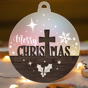 May include: A wooden Christmas ornament shaped like a ball with a white cross, holly leaves, and snowflakes. The ornament has a pink and white ombre effect and the text "Merry Christmas" in white.