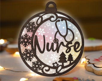 Puede incluir: Un adorno navideño de madera con forma de círculo con un fondo degradado blanco y rosa. El adorno presenta un estetoscopio, copos de nieve, estrellas y la palabra "Enfermera".