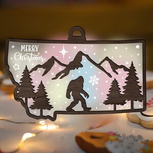 May include: A wooden Christmas ornament shaped like the state of Montana with a silhouette of a Bigfoot walking through a snowy mountain scene. The ornament is decorated with a rainbow gradient and the text "Merry Christmas".