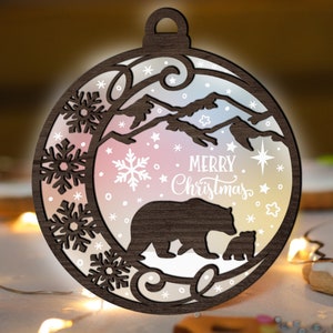 May include: A wooden Christmas ornament shaped like a circle with a brown bear and a cub inside. The ornament has a white background with snowflakes and the text "Merry Christmas".