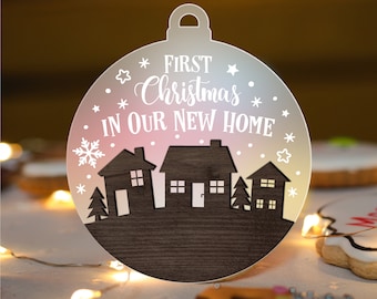 May include: A wooden Christmas ornament shaped like a ball with a white snowflake and stars on a pink and blue ombre background. The ornament says "First Christmas in our new home" with a silhouette of houses and trees.