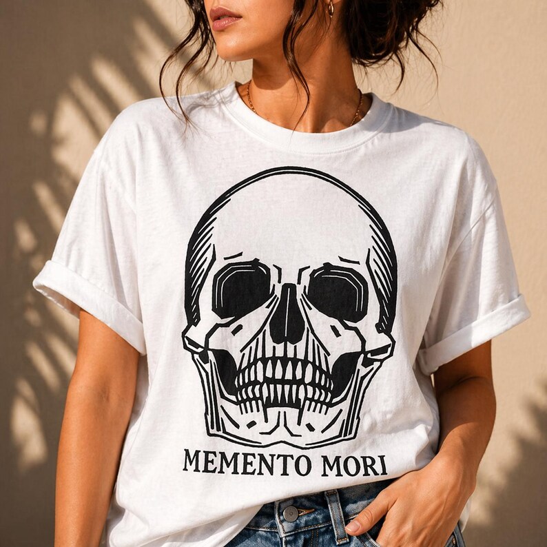 May include: White t-shirt featuring a large black skull graphic. The skull has detailed shading and a prominent jawline. Below the skull, the words "MEMENTO MORI" are printed in a bold, sans-serif font. The shirt has short sleeves and a relaxed fit.