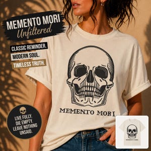 May include: Cream-colored t-shirt featuring a large black skull graphic. The shirt has the words "MEMENTO MORI" and "Unfiltered" printed on it, along with other text. The shirt is a classic reminder of a modern soul and timeless truth.