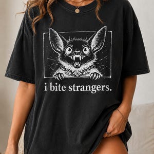 Puede incluir: Una camiseta negra de manga corta con un gráfico blanco de un murciélago con ojos grandes y dientes afilados dentro de un marco rectangular. Debajo de la ilustración, el texto "i bite strangers." aparece en una fuente blanca sencilla.