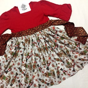 May include: Red and white Christmas dress with a nutcracker print and a brown and gold ribbon. The dress has a long sleeve with a ruffle at the elbow.