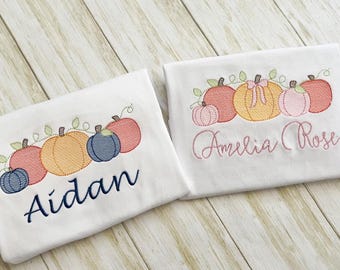Könnte beinhalten: Zwei weiße Hemden mit gestickten Kürbismotiven. Ein Hemd hat den Namen "Aidan" in Marineblau, das andere "Amelia Rose" in Rosa. Die Kürbisse sind in Orange-, Rosa- und Blautönen gehalten.