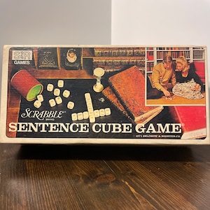 May include: A vintage Scrabble Sentence Cube Game box with a white background and red, black, and white text. The box features a photo of two people playing the game with a wooden table, books, and a sand timer.