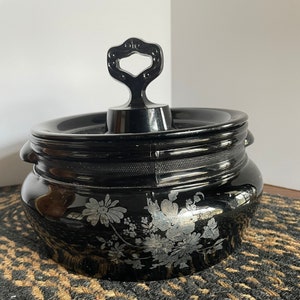 Puede incluir: Un tarro de vidrio negro con un diseño floral plateado y una tapa negra con un asa decorativa.