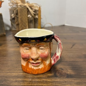 May include: A ceramic mug shaped like a man's face with a red beard and rosy cheeks. The mug has a black rim with yellow accents and a handle with red and white patterns. The mug is on a wooden surface.