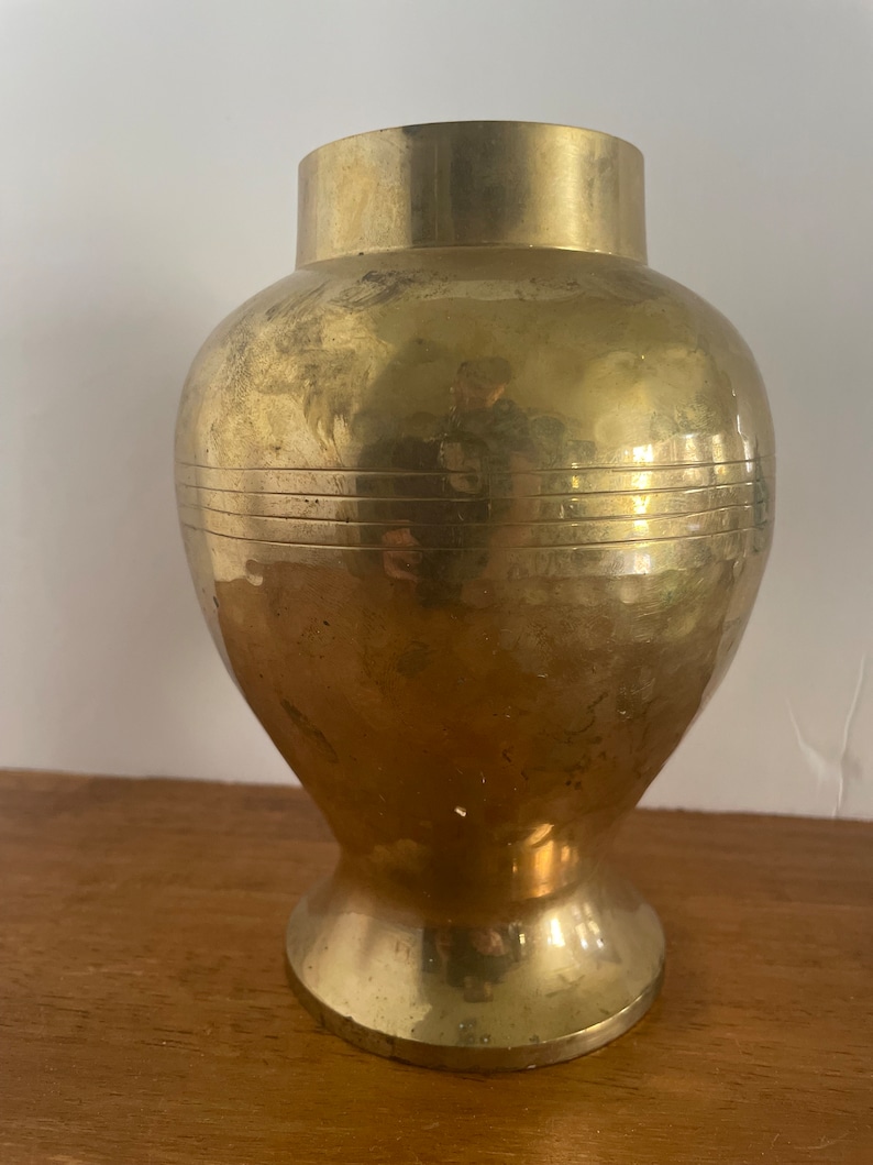 May include: A gold brass vase with a round body and a flared base. The vase has a smooth, polished finish and a subtle, textured pattern.