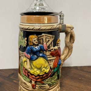 Op de afbeelding: Een keramische bierpul met een reliëfontwerp van een persoon in een blauwe top en gele rok die danst naast een persoon die fluit speelt. De pul heeft een metalen deksel met een reliëfpatroon en een getextureerd handvat dat lijkt op een tak.
