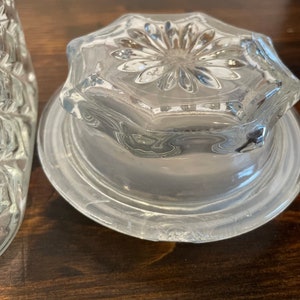 Op de afbeelding: Een deksel van helder glas met een decoratief bloemmotief aan de bovenkant. Het deksel heeft een verhoogde rand en een glad, gepolijst oppervlak.