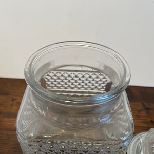 Op de afbeelding: Een vierkante glazen pot met een transparante kleur en een textuur van een diamantpatroon. De pot heeft een deksel.