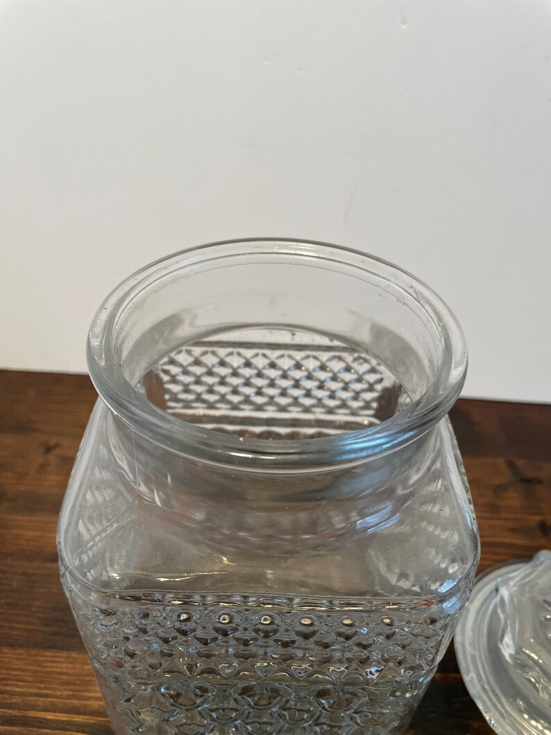 Op de afbeelding: Een vierkante glazen pot met een transparante kleur en een textuur van een diamantpatroon. De pot heeft een deksel.