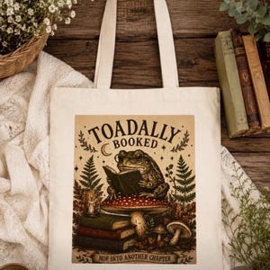 May include: A cream-colored canvas tote bag featuring a graphic print of a toad wearing glasses and reading a book while sitting on a mushroom. The text reads TOADALLY BOOKED and HOP INTO ANOTHER CHAPTER, surrounded by ferns and stacked books.