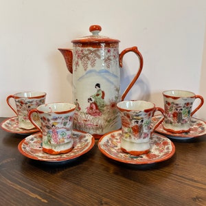 Puede incluir: Un juego de té japonés vintage con una tetera blanca y cuatro tazas y platillos. La tetera y las tazas están decoradas con un patrón floral y una escena de mujeres con ropa tradicional japonesa. El juego está en excelente estado.