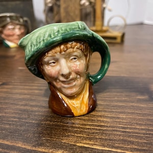 May include: A ceramic figurine of a smiling man wearing a green hat and brown jacket. The figurine has a handle and spout, suggesting it may be a small pitcher or creamer. The face is detailed with painted features.