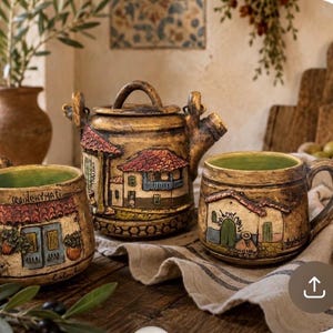 Puede incluir: Un juego de té de cerámica que incluye una tetera y dos tazas con un acabado marrón texturizado y desgastado. Cada pieza presenta un relieve pintado a mano de una casa con techo rojo y el texto Quinchamali, con un esmalte interior de color verde.