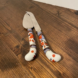 May include: Two silver cheese knives with floral patterned handles. The handles are white with red, blue, and gold accents.