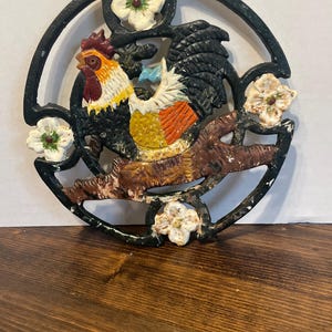 Könnte beinhalten: Ein gusseiserner Untersetzer mit einem Hahn-Design. Der Hahn ist in Schwarz, Gelb, Rot, Orange und Weiß bemalt. Der Untersetzer ist mit weißen Blumen mit grünen Blättern und roten Zentren verziert.