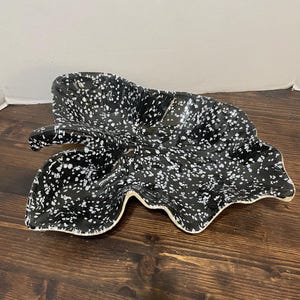 May include: A leaf-shaped ceramic serving dish featuring a black glaze with a white speckled pattern. The dish has a distinct stem-like handle on one side and an irregular, wavy edge that mimics the natural form of a large foliage leaf.