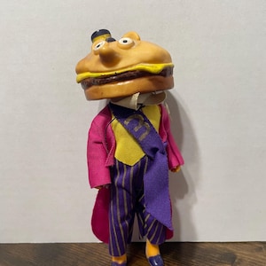 May include: A vintage toy figure of a hamburger character wearing a purple suit with gold stripes and a yellow vest. The figure has a large hamburger head with a sesame seed bun, lettuce, tomato, and a meat patty.