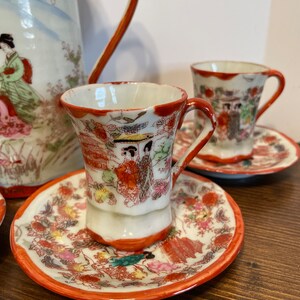 May include: A porcelain tea set featuring a pitcher and two cups with matching saucers. The white ceramic is decorated with orange rims and detailed illustrations of figures in traditional clothing, pagodas, and floral patterns in various colors.