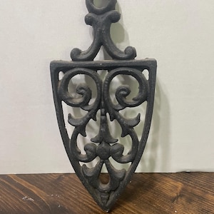 May include: Black cast iron trivet with an ornate, openwork design. The trivet has a shield shape with a handle at the top. The decorative pattern includes scrolling lines and floral elements. The trivet is designed to protect surfaces from hot cookware.