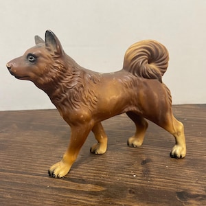 Pode incluir: Uma estatueta de cão castanha com cauda enrolada, orelhas pontiagudas e detalhes de pelo texturizado. A figura canina está de perfil, apresentando um focinho castanho escuro, nariz preto e patas castanho-claras com acabamento mate.