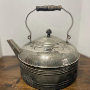 Peut inclure: Bouilloire ancienne en métal argenté avec une poignée incurvée et une poignée en bois. La bouilloire a un bec et un couvercle avec un petit bouton. La bouilloire repose sur une surface en bois.