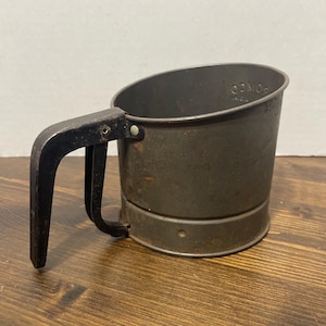 May include: Vintage metal flour sifter with a dark handle and a cylindrical body. The sifter has a dark, aged appearance, suggesting it is an antique kitchen tool. The handle is black and attached to the side of the sifter.