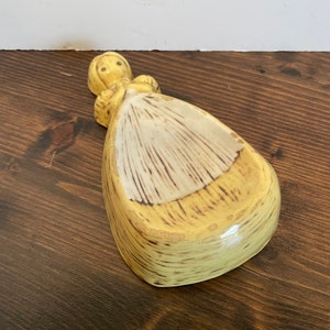 May include: A yellow ceramic spoon rest shaped like a woman in a dress. The woman has a white dress with brown stripes and a yellow bodice.