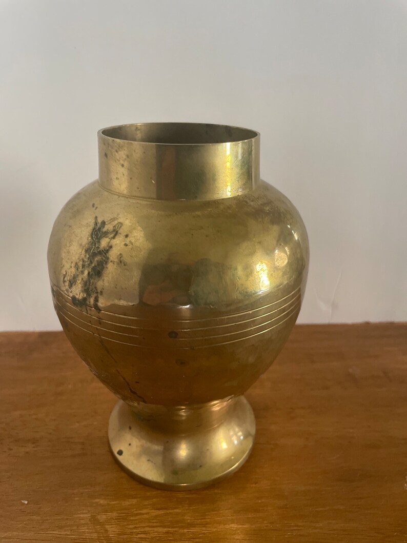 May include: A gold brass vase with a round body and a flared base. The vase has a smooth, polished finish and a subtle, textured pattern.