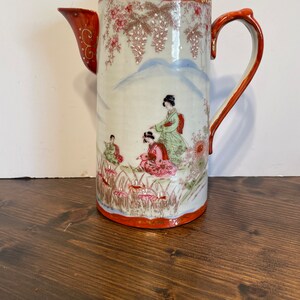 May include: A cylindrical porcelain coffee pot featuring a hand-painted scene of figures in traditional attire amidst floral motifs and mountain silhouettes. The vessel has a red lid, a curved handle, and a spout with gold-painted accents.