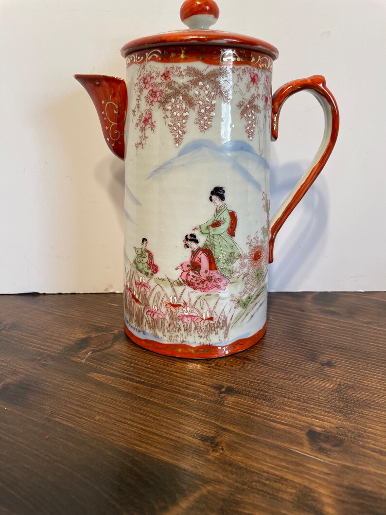 May include: A cylindrical porcelain coffee pot featuring a hand-painted scene of figures in traditional attire amidst floral motifs and mountain silhouettes. The vessel has a red lid, a curved handle, and a spout with gold-painted accents.