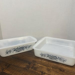 May include: Two white milk glass baking dishes featuring a dark blue floral and leaf pattern along the exterior sides. One dish is rectangular and deep, while the other is square and shallow, both with smooth, glossy finishes and clean edges.