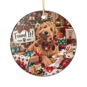 May include: A round ceramic Christmas ornament featuring a golden doodle dog holding a plush reindeer toy in its mouth. The dog wears a red and black plaid bandana with a white paw print. A sign reads Found It! with paw prints and a plaid bow.