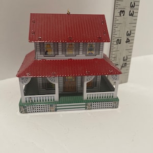 May include: A white and red tin house ornament with a green porch and a red roof. The house has windows with curtains and a door. The ornament has a gold hanger.