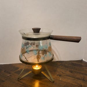 Puede incluir: Una tetera calentadora de cristal transparente con mango y tapa de madera, decorada con un patrón de piñas y agujas azules. Se apoya sobre un soporte de trípode de metal dorado con una vela de té encendida debajo para dar calor.