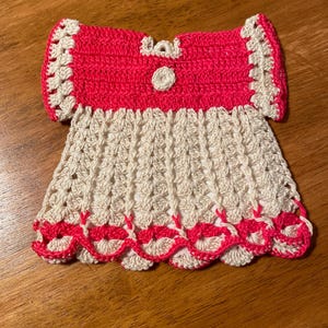 May include: A crocheted doll dress with a pink bodice and a cream-colored skirt. The dress has a scalloped hem and a white button.