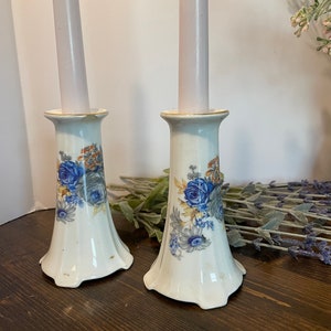 May include: A pair of white ceramic candle holders with a floral design featuring blue roses and yellow accents. The holders have a fluted base and a smooth, cylindrical top.