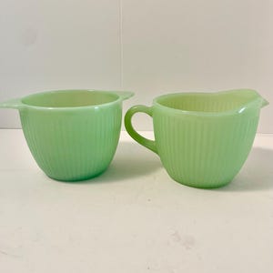 Puede incluir: Dos cuencos de vidrio verde jade. Uno tiene dos asas y el otro tiene un asa y un pico. Ambos cuencos tienen detalles verticales acanalados. Probablemente sean utensilios de cocina antiguos.