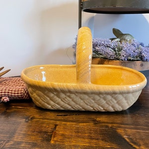 Puede incluir: Una cesta de cerámica con un diseño tejido en color marrón claro. La cesta tiene un asa y es perfecta para guardar objetos pequeños.